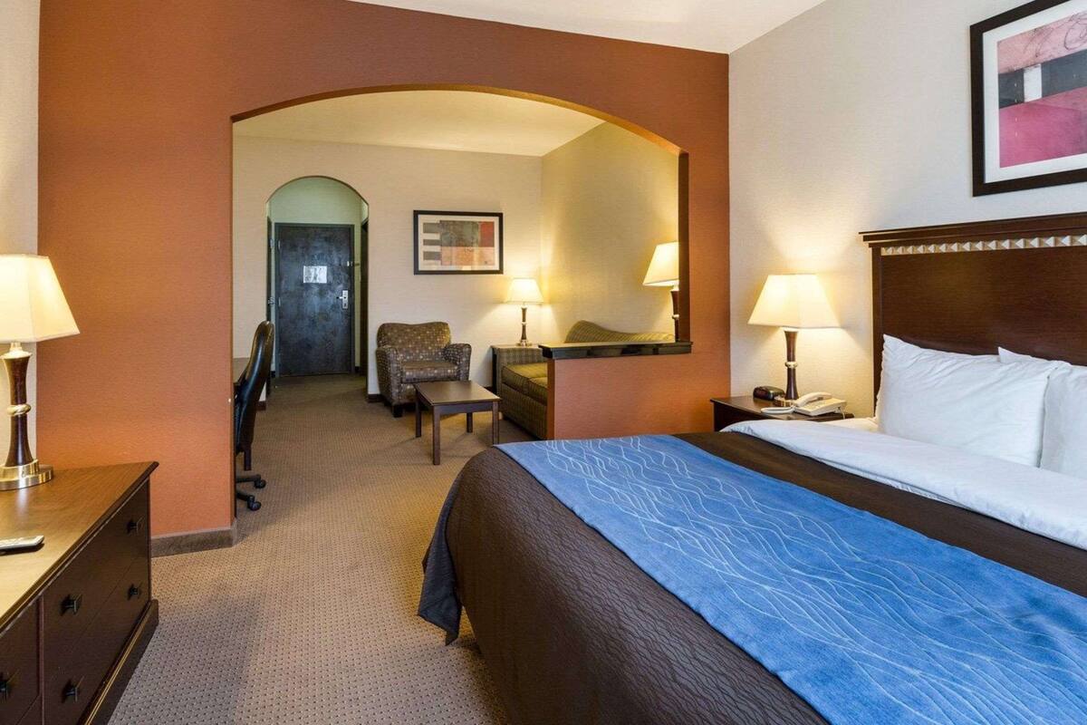 Ein Hotelzimmer mit einem Bett, zwei Stühlen, einem Schreibtisch, einer Kommode, einem Spiegel und einer Tür zu einem anderen Raum.