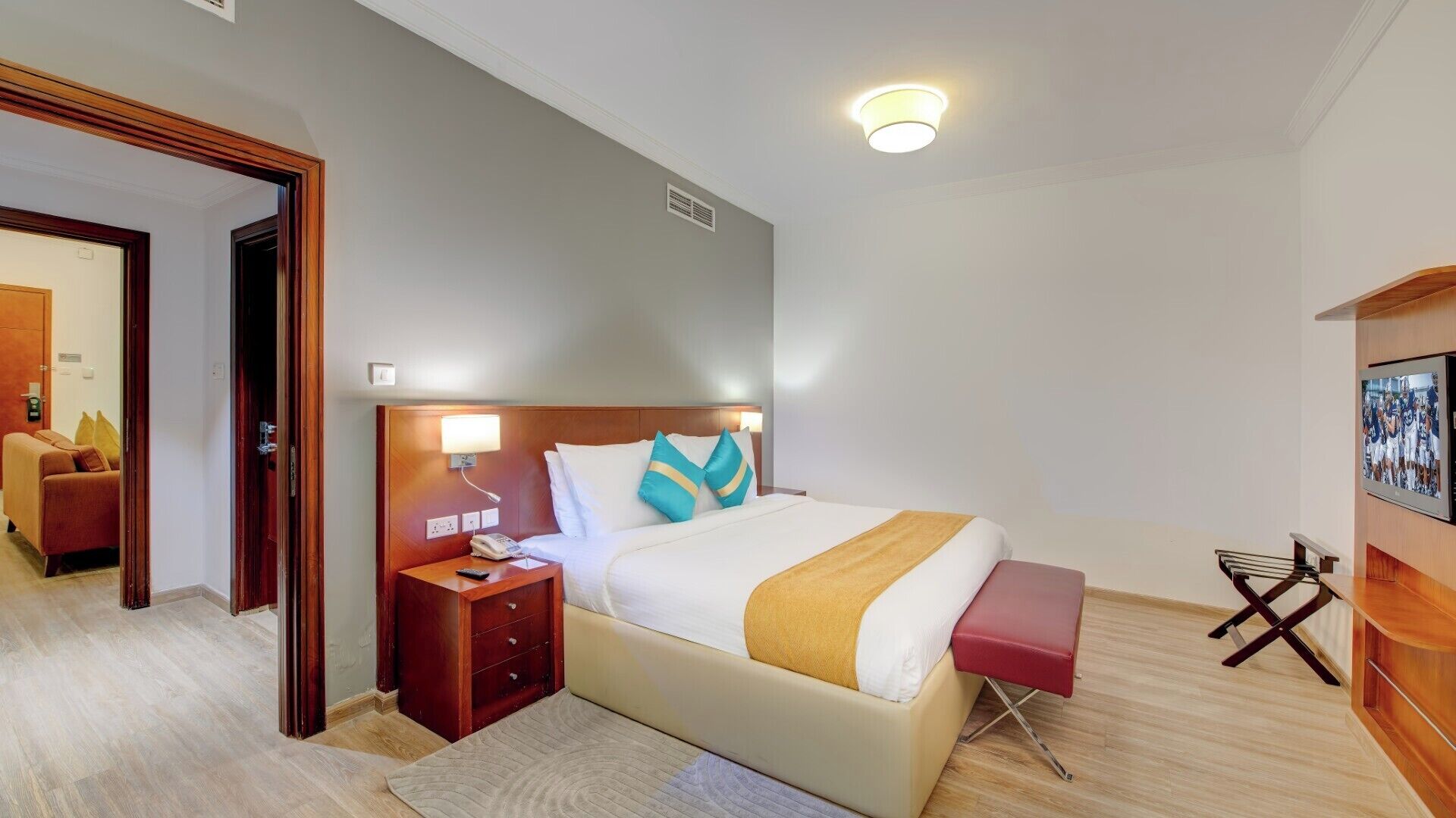 A hotel room with a bed, bedside tables, a bench, a TV, and a door leading to another room.