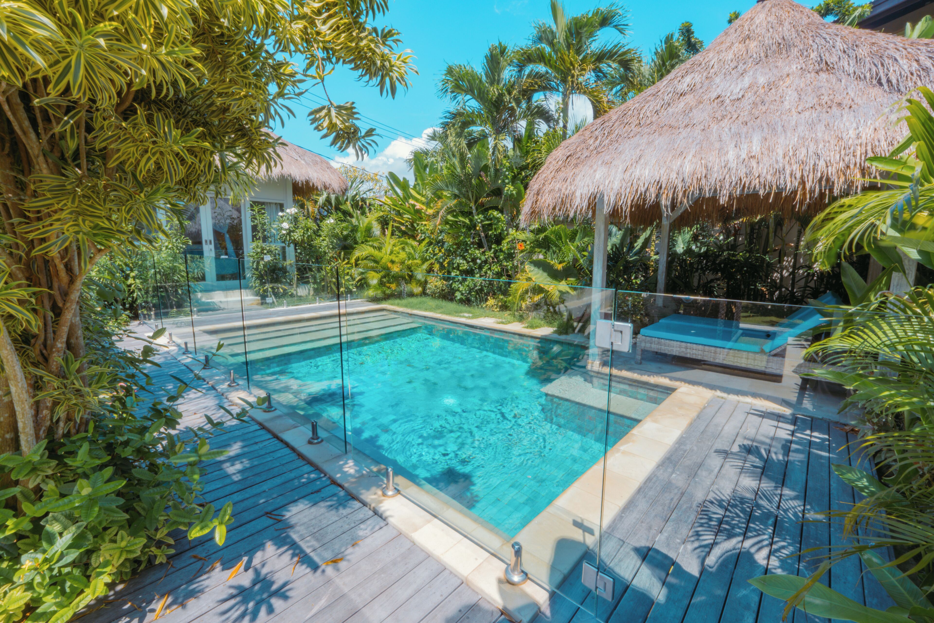 A pool with a glass barrier, surrounded by tropical vegetation and thatched umbrellas.