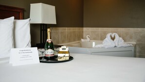 A hotel room with a bed, a bedside table with a champagne bottle, a glass, and a card. There is a bathtub with white swan decorations in the background. - GrandStay Hotel & Suites Perham (Perham)