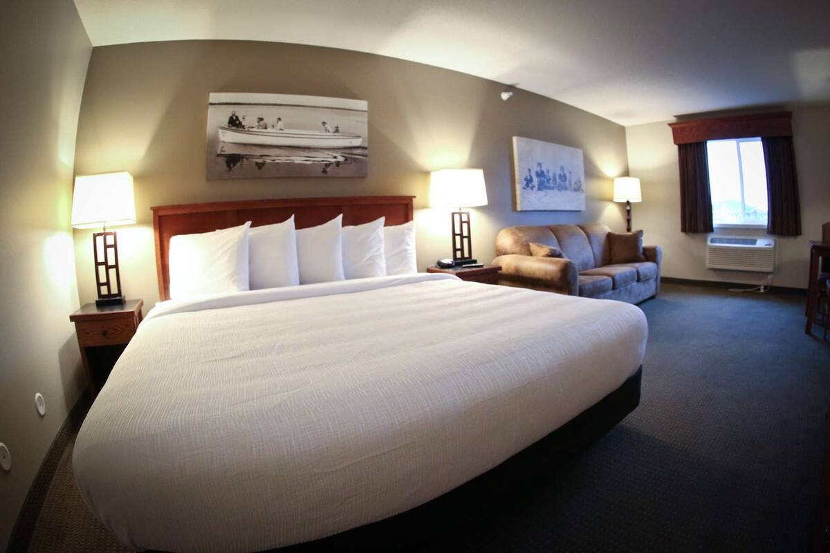 A large bed with white linens, two bedside tables with lamps, a brown leather sofa, a wall-mounted artwork, and a window with curtains.