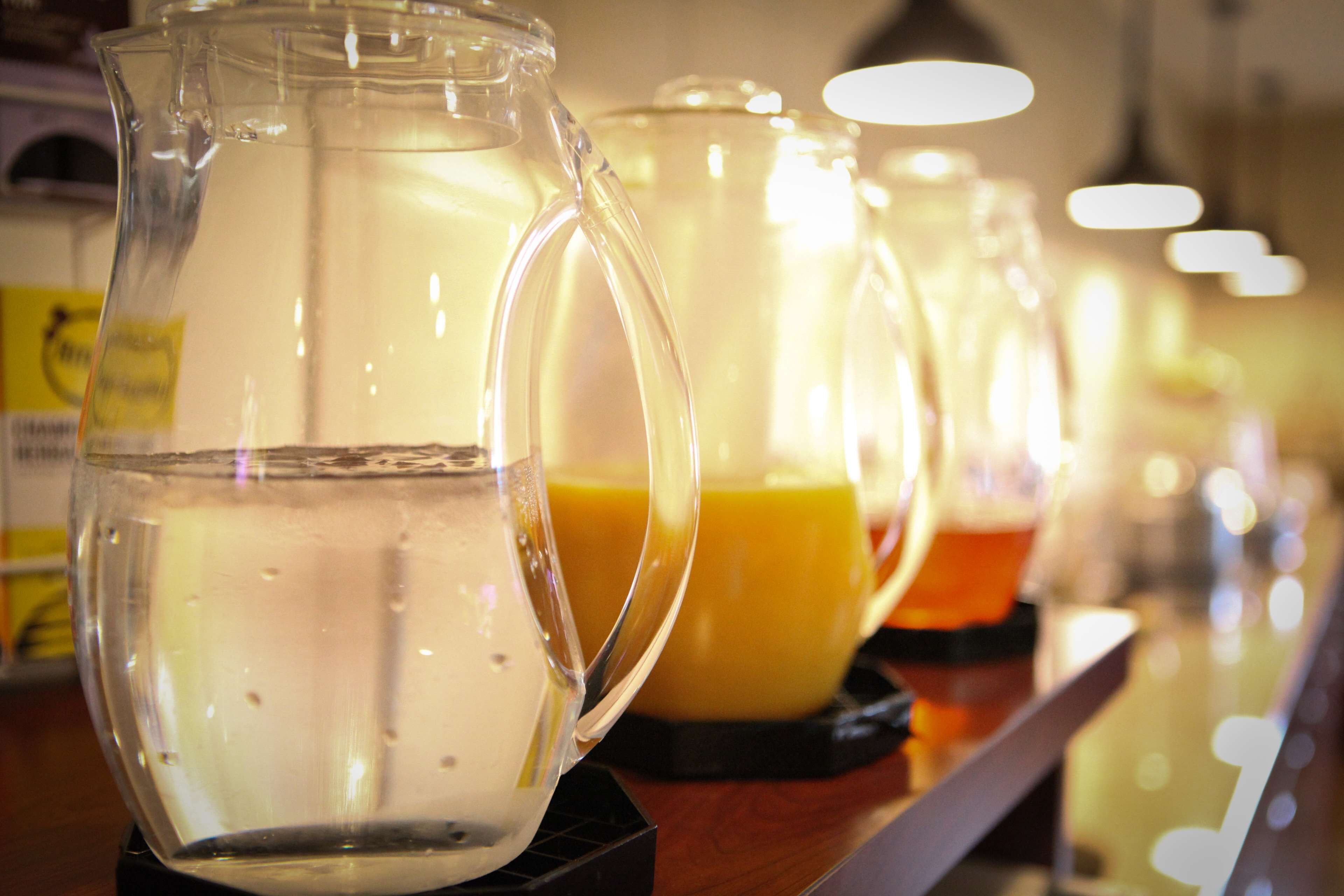 Une rangée de carafes en verre transparent, remplies d’eau et de jus d’orange, est posée sur une surface en bois.