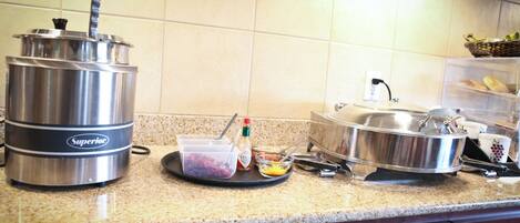 A kitchen counter with a food warmer, a plastic container with condiments, and a basket of bread.