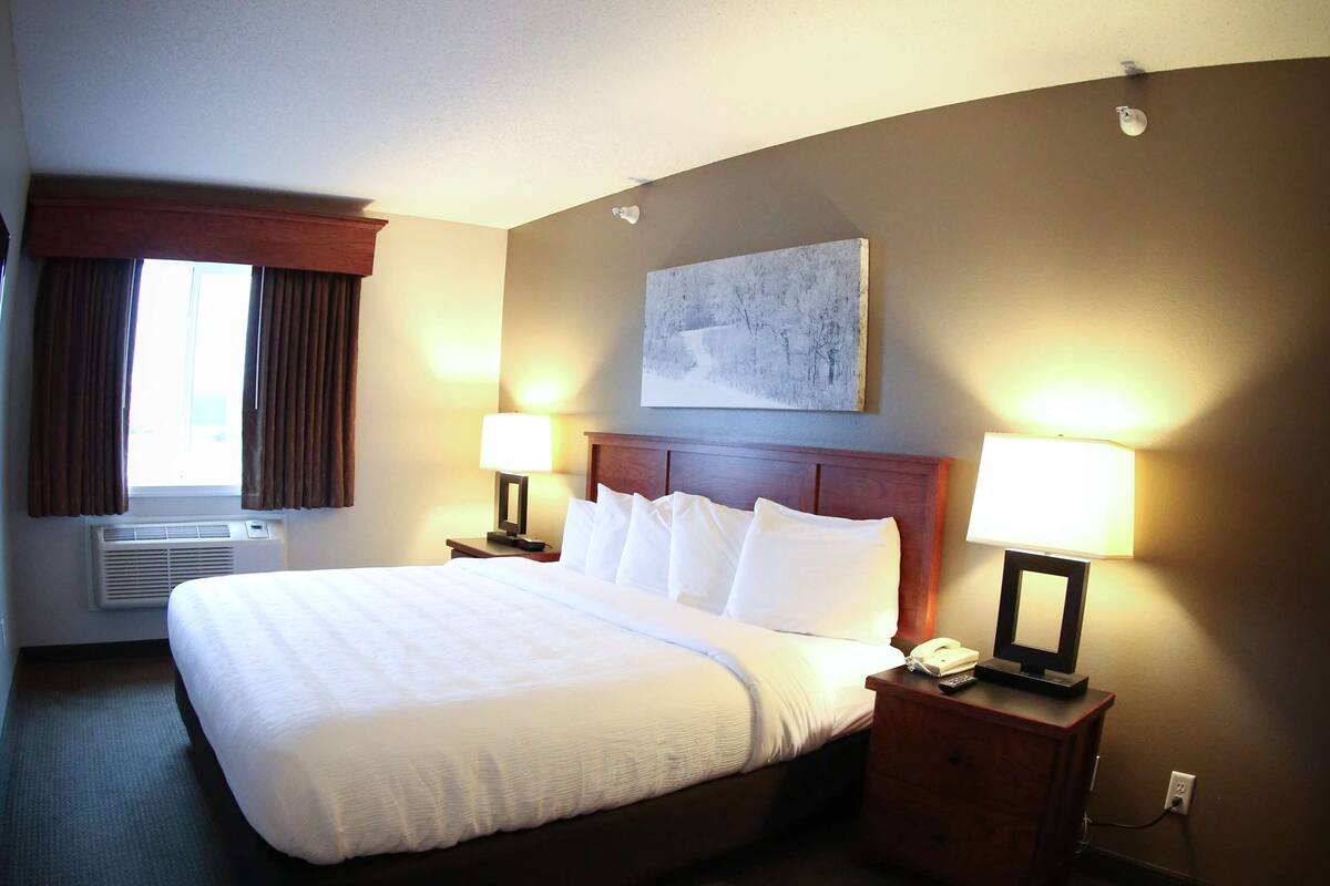 A hotel room with a large bed, two bedside tables with lamps, a window with curtains, an air conditioning unit, and a painting on the wall.