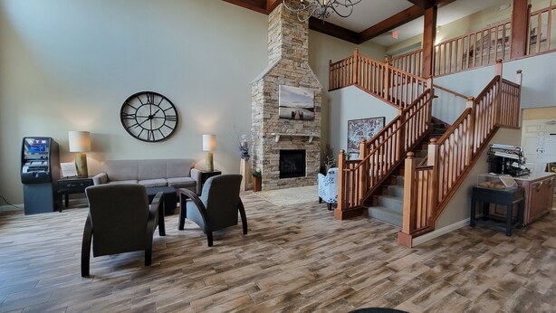 A spacious living area with a stone fireplace, a wooden staircase, and a large wall clock. - GrandStay Hotel & Suites Perham (Perham)