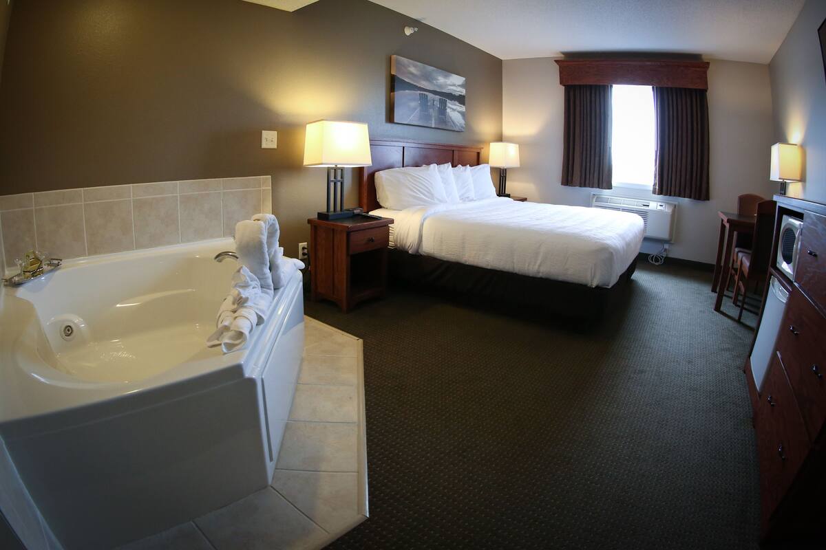 A hotel room with a large bathtub, a bed with white linens, two bedside tables with lamps, a window with curtains, and a painting on the wall.