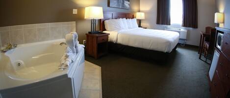 A hotel room with a large bathtub, a bed with white linens, two bedside tables with lamps, a window with curtains, and a painting on the wall.