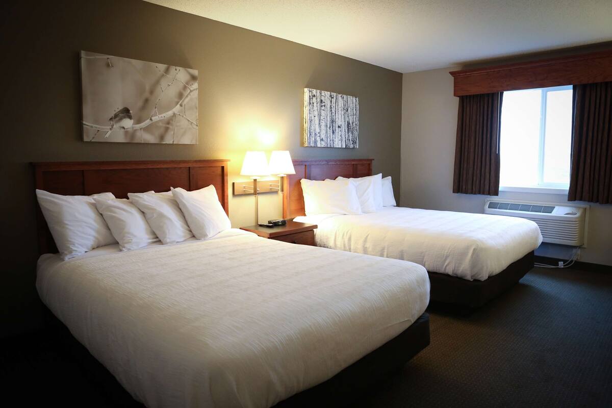 A hotel room with two beds, a nightstand with a lamp, a wall-mounted artwork, and a window with curtains.