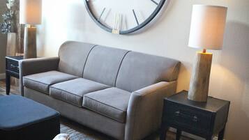 A living room with a large wall clock, a sofa, two side tables with lamps, and a patterned ottoman.