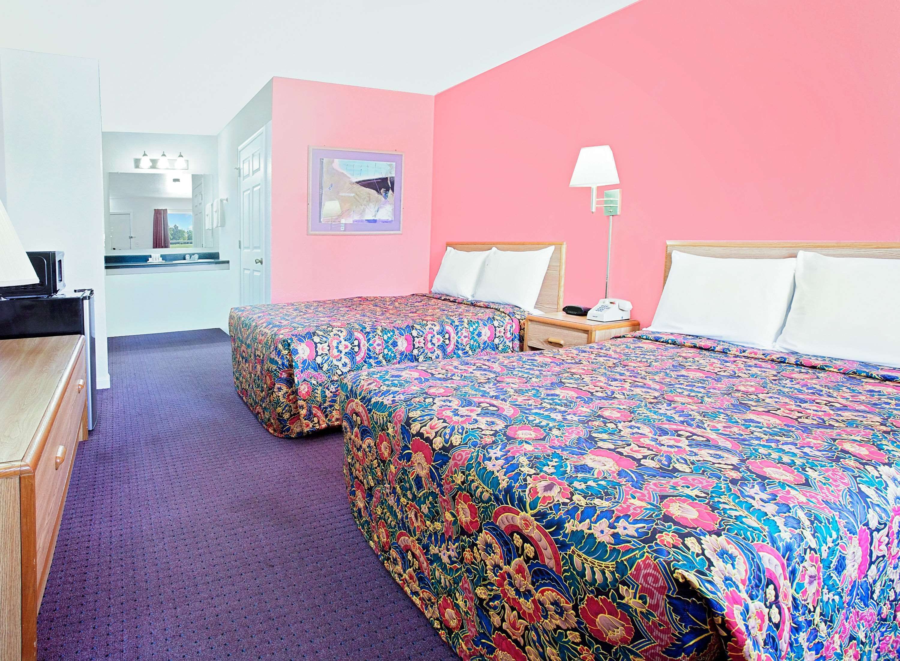 Habitación de hotel con dos camas, un escritorio, una silla, una lámpara y un baño a la vista a través de una puerta abierta.