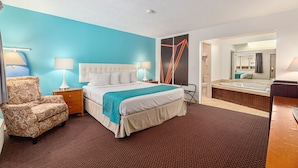 A hotel room with a large bed, a chair, a nightstand, a lamp, a bathroom with a bathtub, and a mirror.