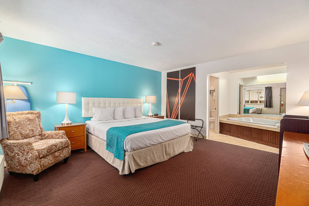 Ein Hotelzimmer mit einem großen Bett, einem Sessel, einem Nachttisch, einer Lampe und einem Badezimmer mit Badewanne und Spiegel.