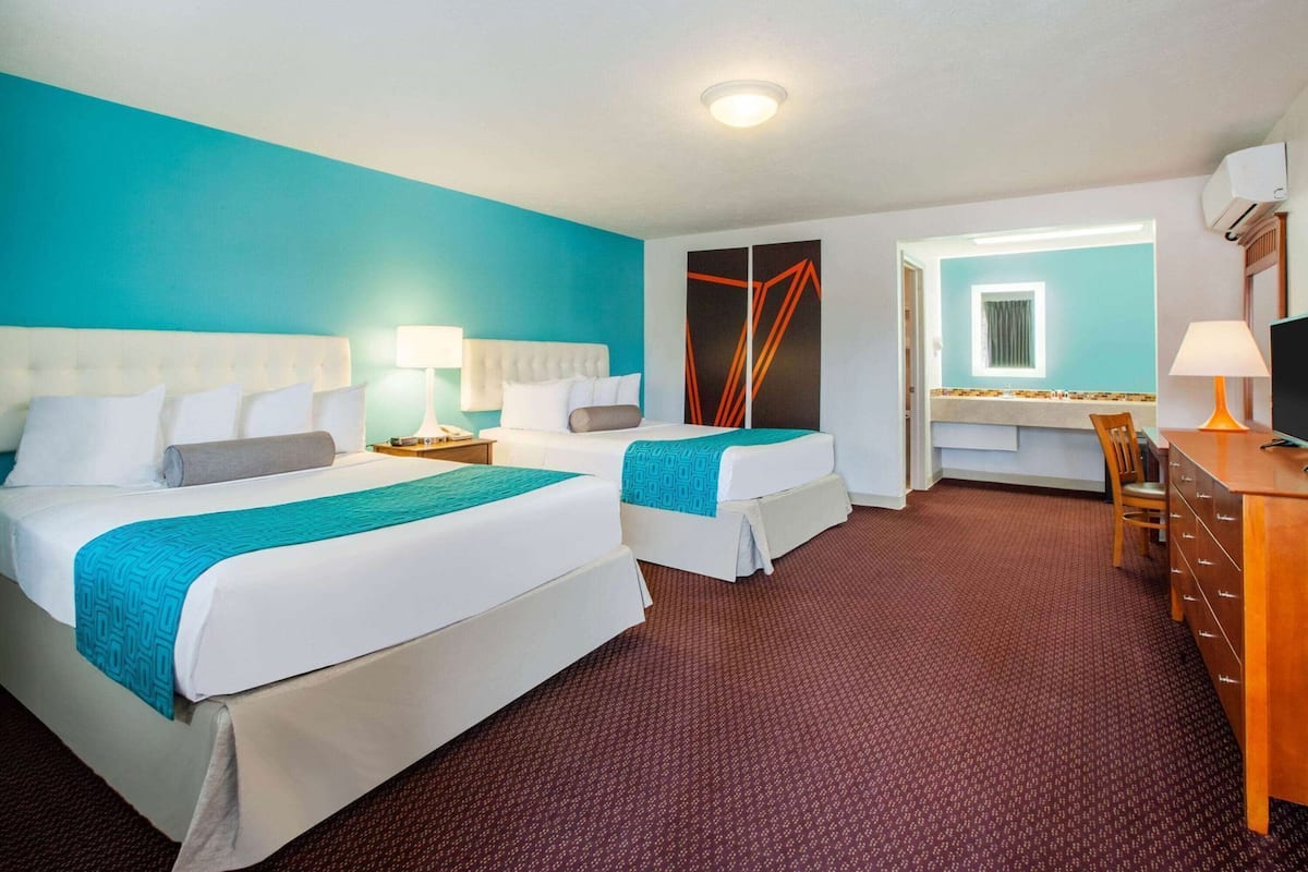 Ein Hotelzimmer mit zwei Betten, einem Schreibtisch, einem Stuhl und einem durch eine offene Tür einsehbarer Badezimmer.