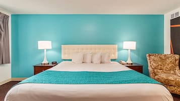A hotel room with a large bed, two bedside tables with lamps, a patterned throw blanket, and a patterned armchair.