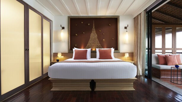 A large bed with white linens and two red pillows, flanked by nightstands with lamps, in a room with wooden flooring and a large window. - Sukhothai Heritage Resort (Sawankhalok)