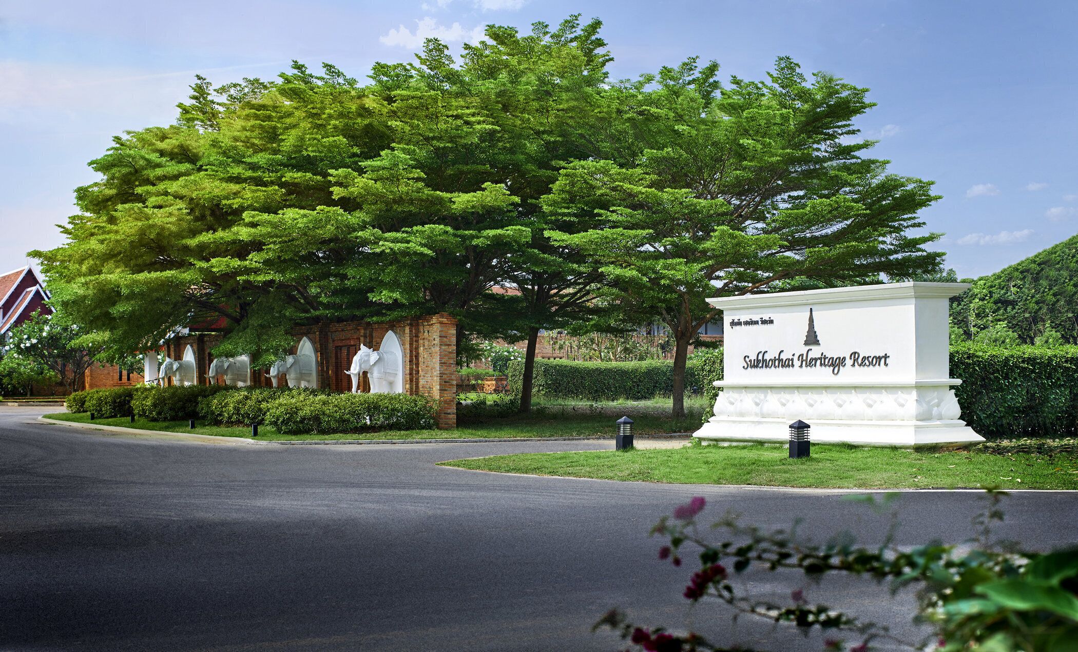 A sign for Sukothai Heritage Resort with a large tree and a brick building in the background.