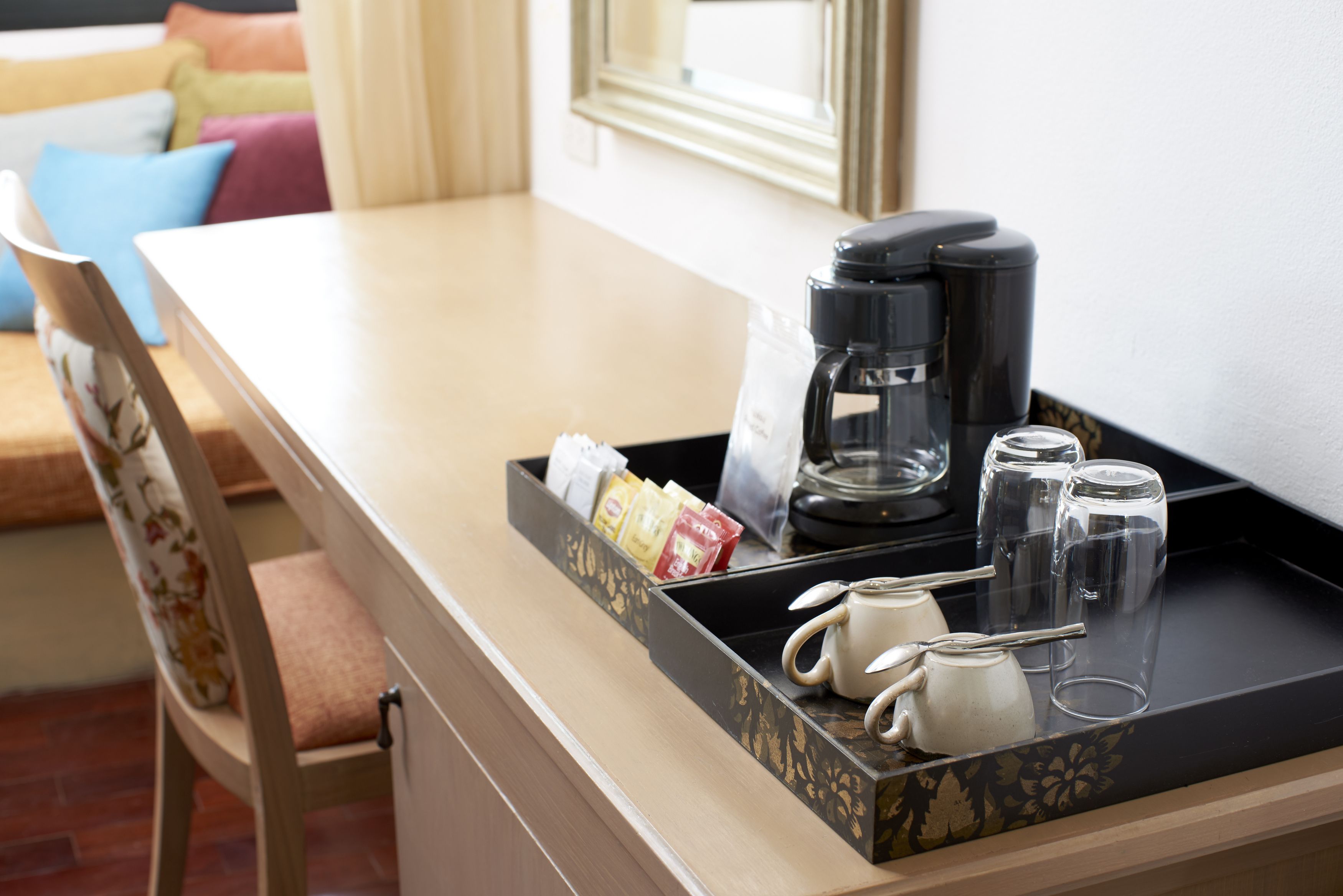 Een hotelkamer met een houten bureau, een koffiezetapparaat, een dienblad met kopjes en een theepot, en een stoel met bloemenmotief.