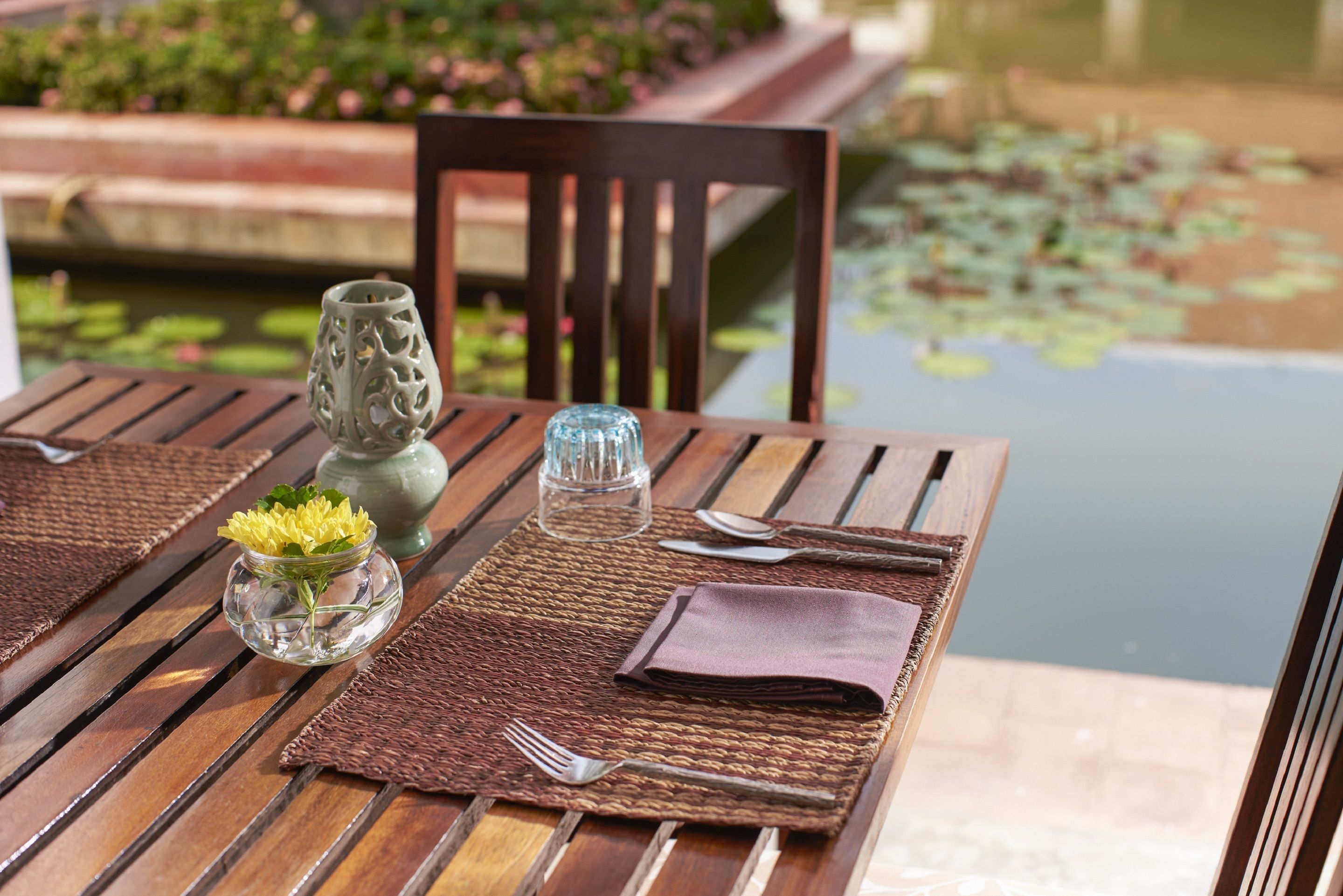 Una mesa de madera con un posavasos tejido, un florero de vidrio con flores amarillas, un vaso transparente y una servilleta doblada.