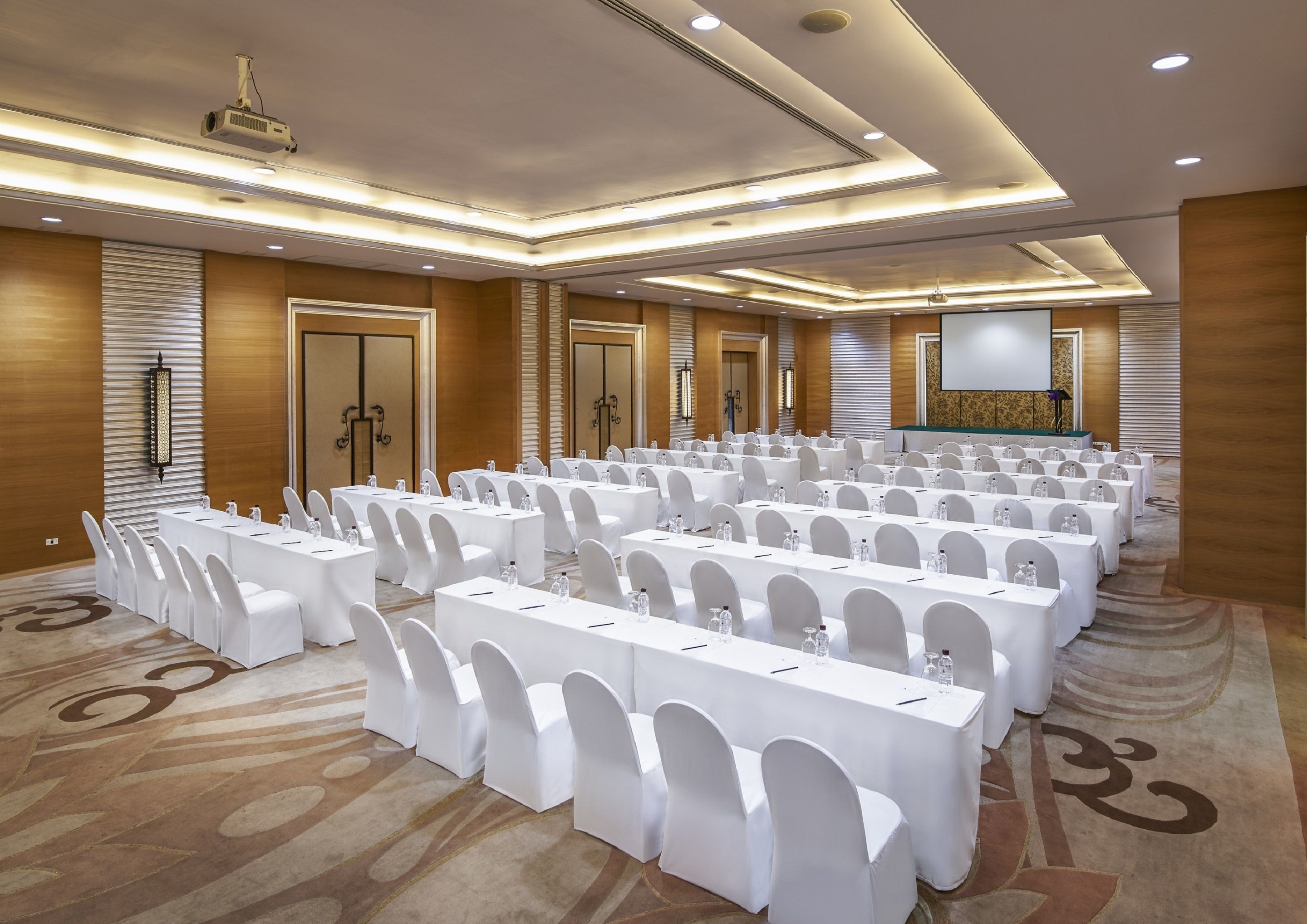 Une salle de conférence avec des rangées de tables et de chaises blanches, un projecteur et un grand écran.