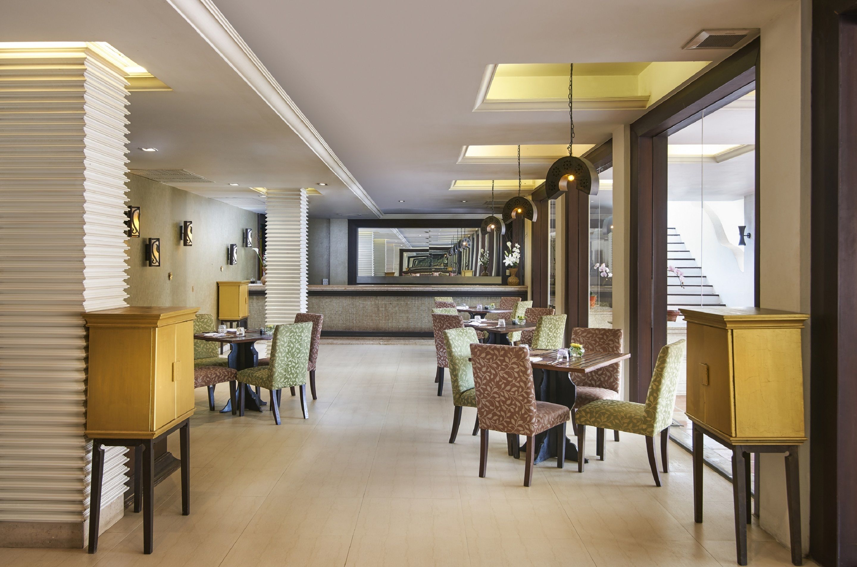 A spacious hotel dining area with multiple tables and chairs, a central bar, and large windows providing natural light.