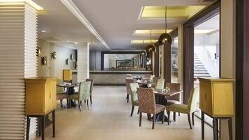 A spacious hotel dining area with multiple tables and chairs, a central bar, and large windows providing natural light.