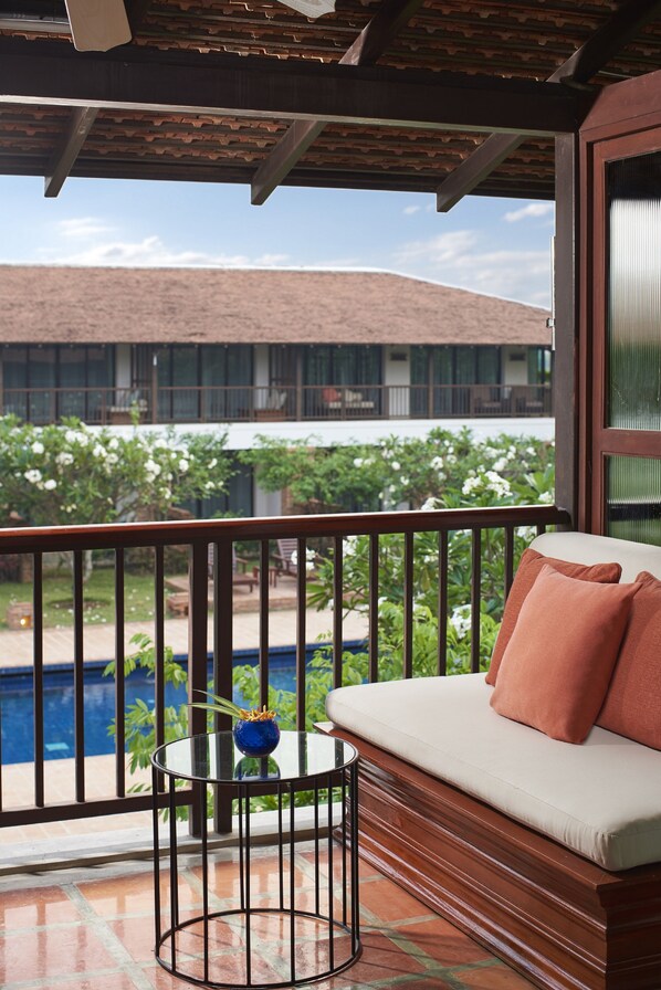 A balcony with a sofa and a small table, overlooking a swimming pool and a building with a red roof.