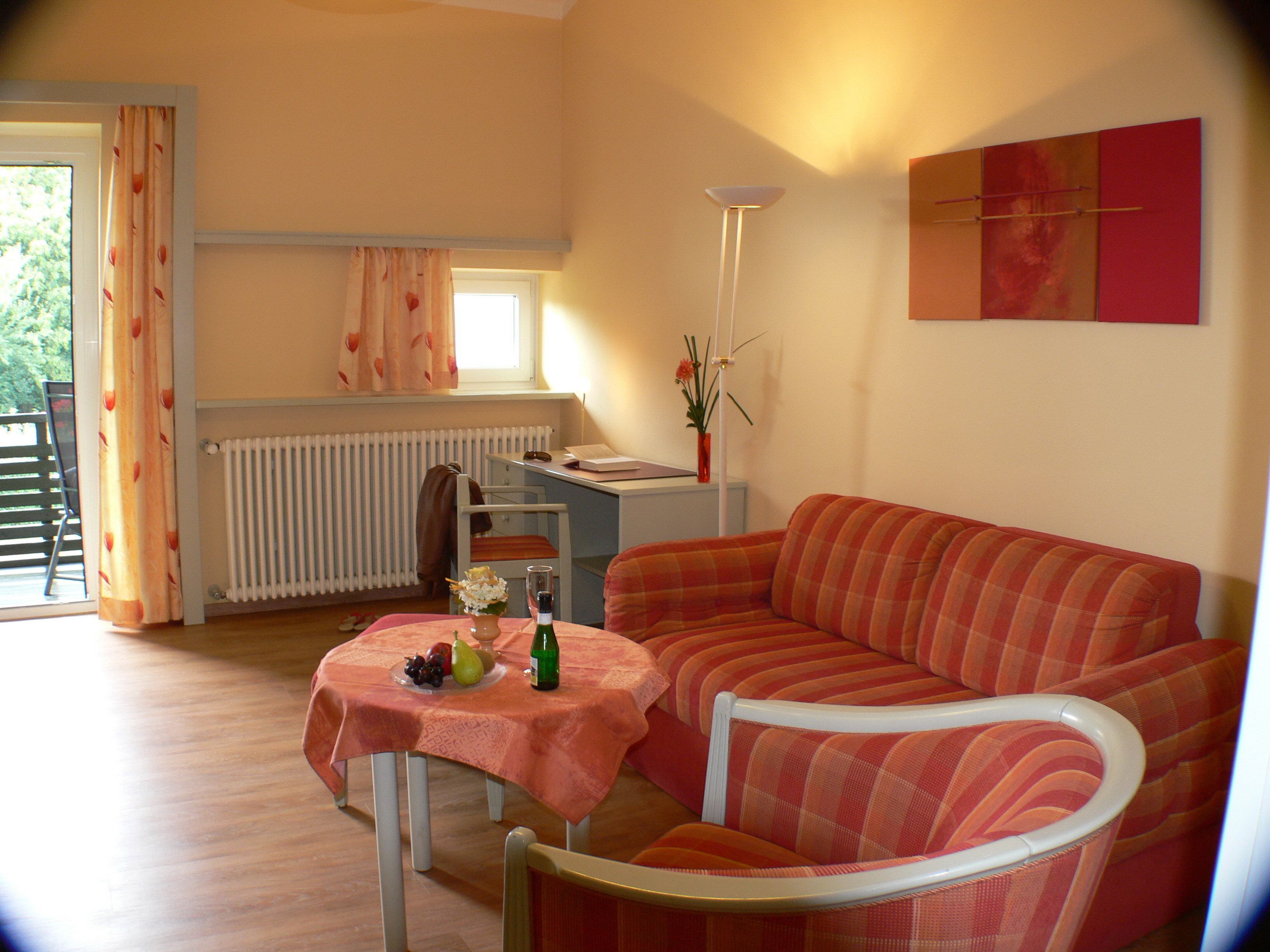 A cozy living room with a sofa, a dining table with a tablecloth, a radiator, a window with curtains, and a floor lamp.
