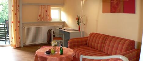 A cozy living room with a sofa, a dining table with a tablecloth, a radiator, a window with curtains, and a floor lamp.