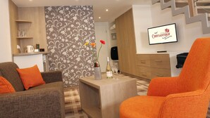 A modern living room with a sofa, armchair, and a small table with a bottle and glasses. A TV is mounted on the wall, and a staircase is visible in the background.
