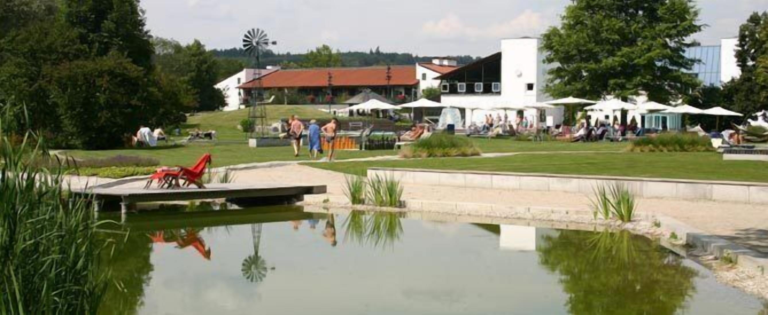 A lakeside setting with a windmill, a small dock, and people relaxing by the water.