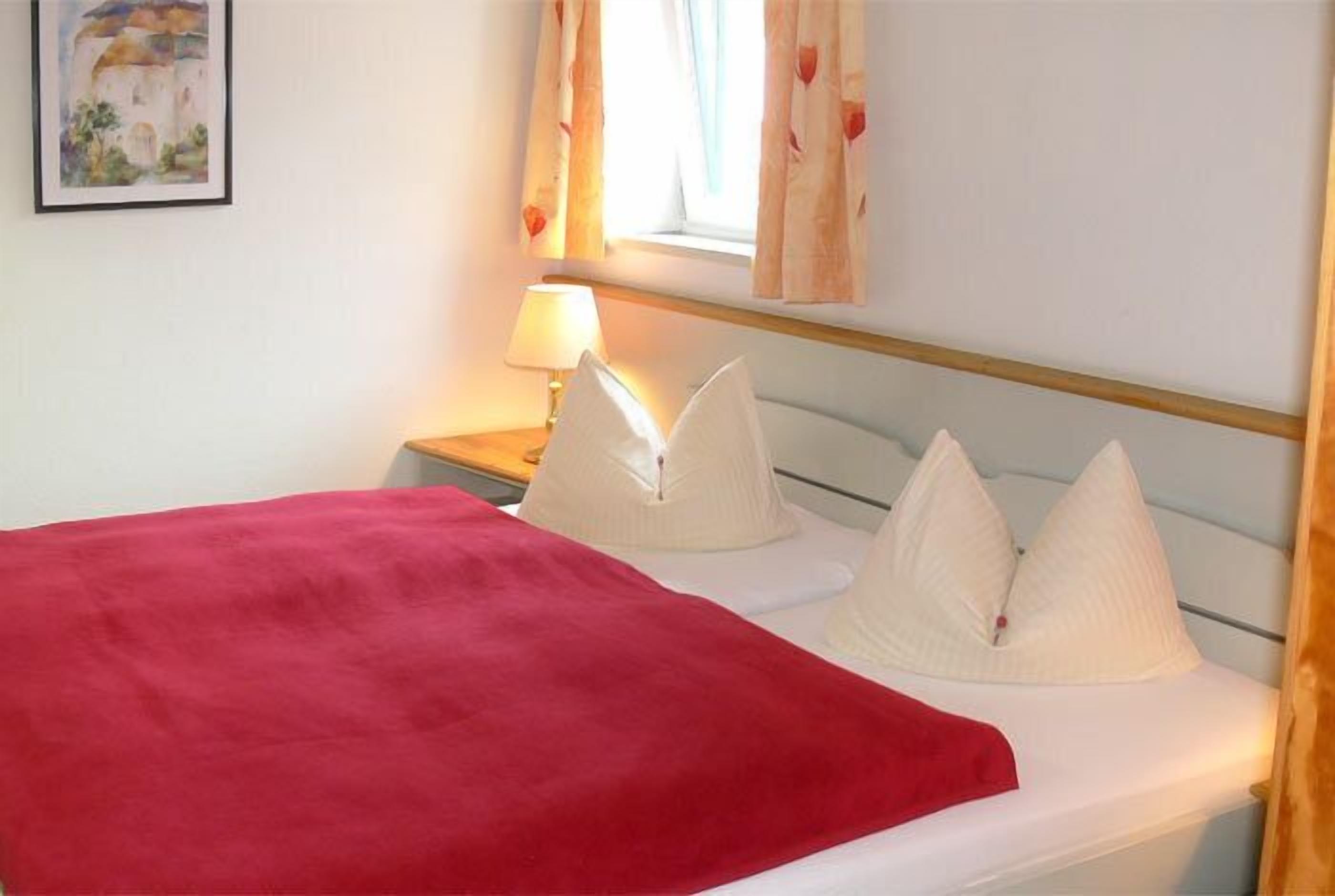 Een bed met een rode deken, witte kussens en een nachtkastje met een lamp.