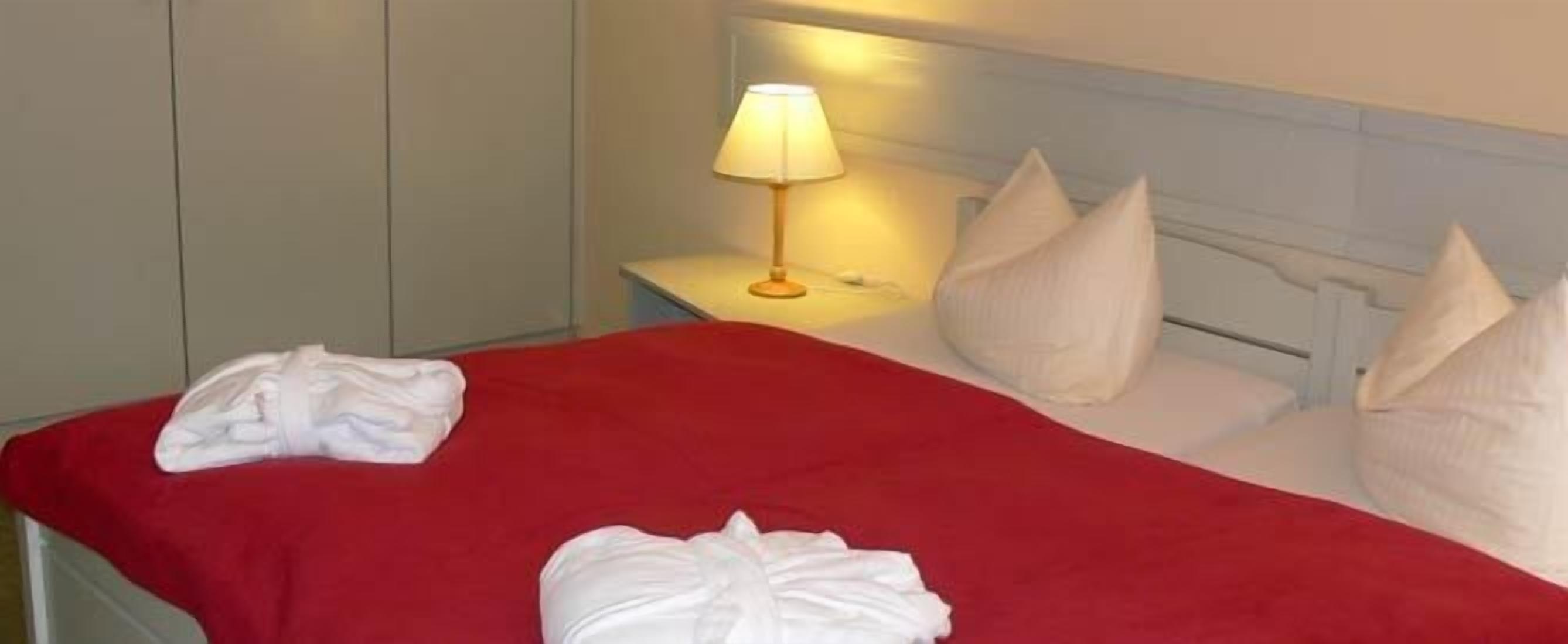 Une chambre d’hôtel avec un lit, des oreillers blancs, une couverture rouge et une lampe de chevet.