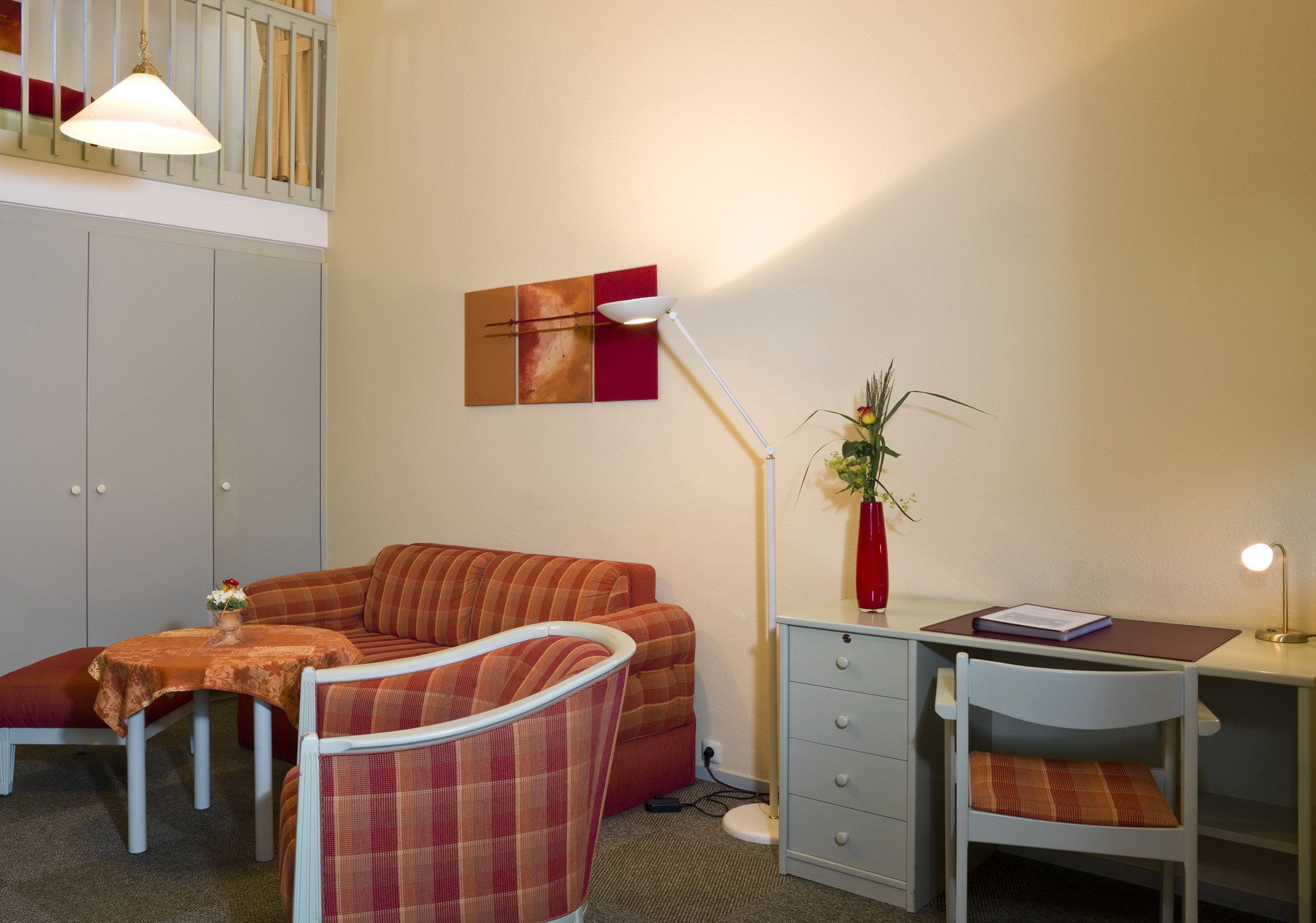 Ein gemütliches Zimmer mit einer Couch, einem kleinen Tisch mit einer Vase, einem Schreibtisch mit einer Lampe und einem Wandbild.