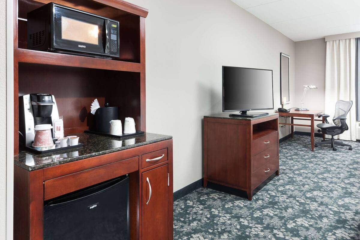A hotel room with a kitchenette, a flat-screen TV, a desk with a chair, and a patterned carpet.