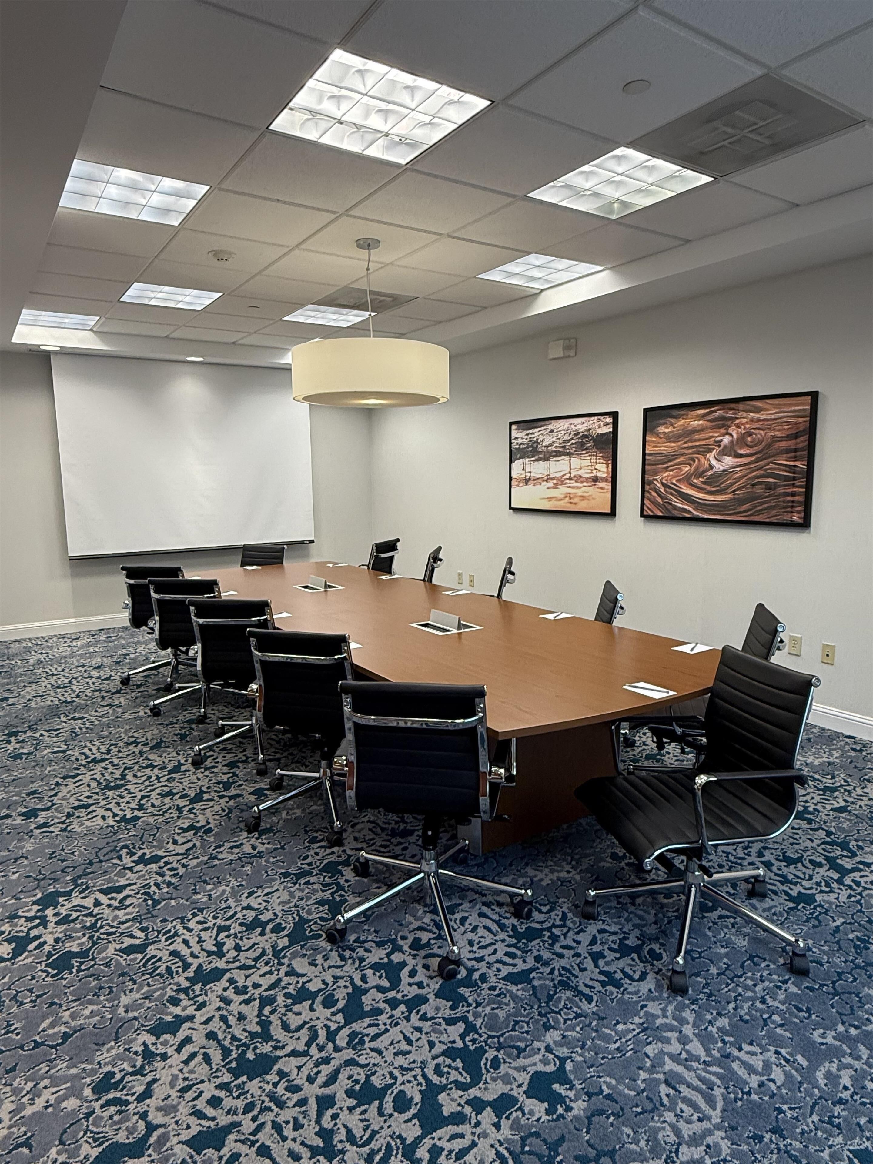 Un salón de conferencias con una gran mesa, sillas, una pantalla de proyector y dos cuadros en la pared.