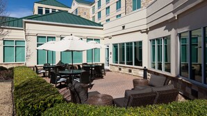 A hotel patio with outdoor seating, large windows, and a brick pathway. - Hilton Garden Inn Frisco (Frisco)