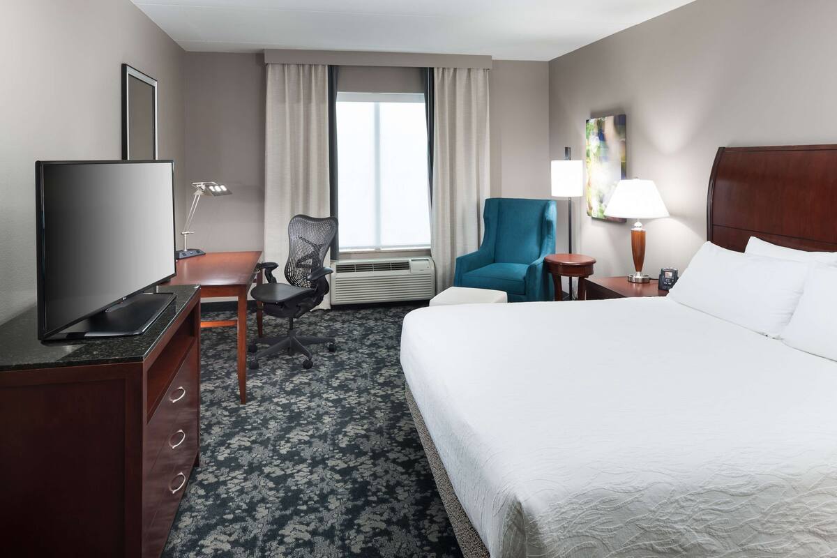 A hotel room with a bed, a desk with a TV, a chair, a lamp, and a window with curtains.