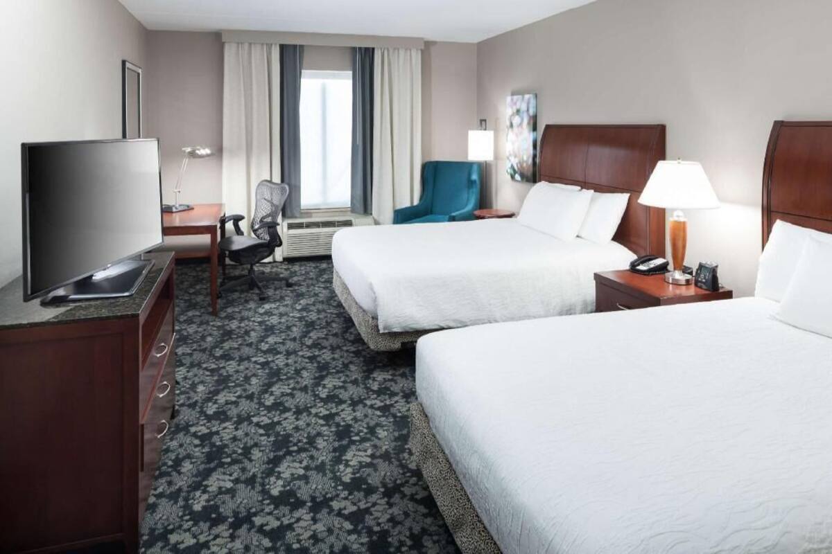 A hotel room with two beds, a desk with a TV, a chair, a lamp, and a window with curtains.