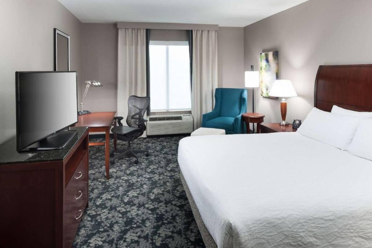 A hotel room with a bed, a desk with a TV, a chair, a lamp, and a window with curtains.