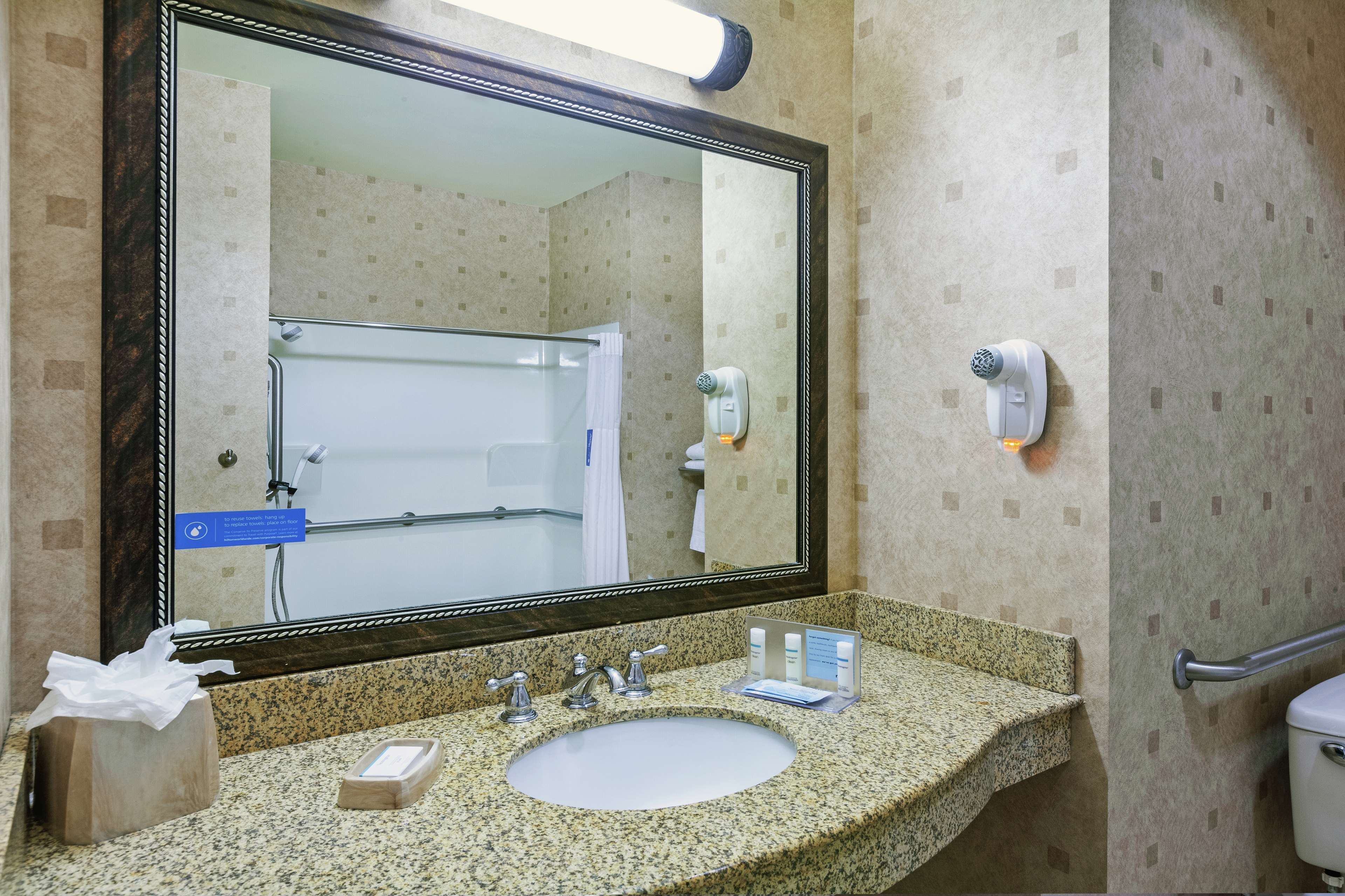 A bathroom with a granite countertop, a large mirror, a sink, a towel dispenser, and a hand dryer.