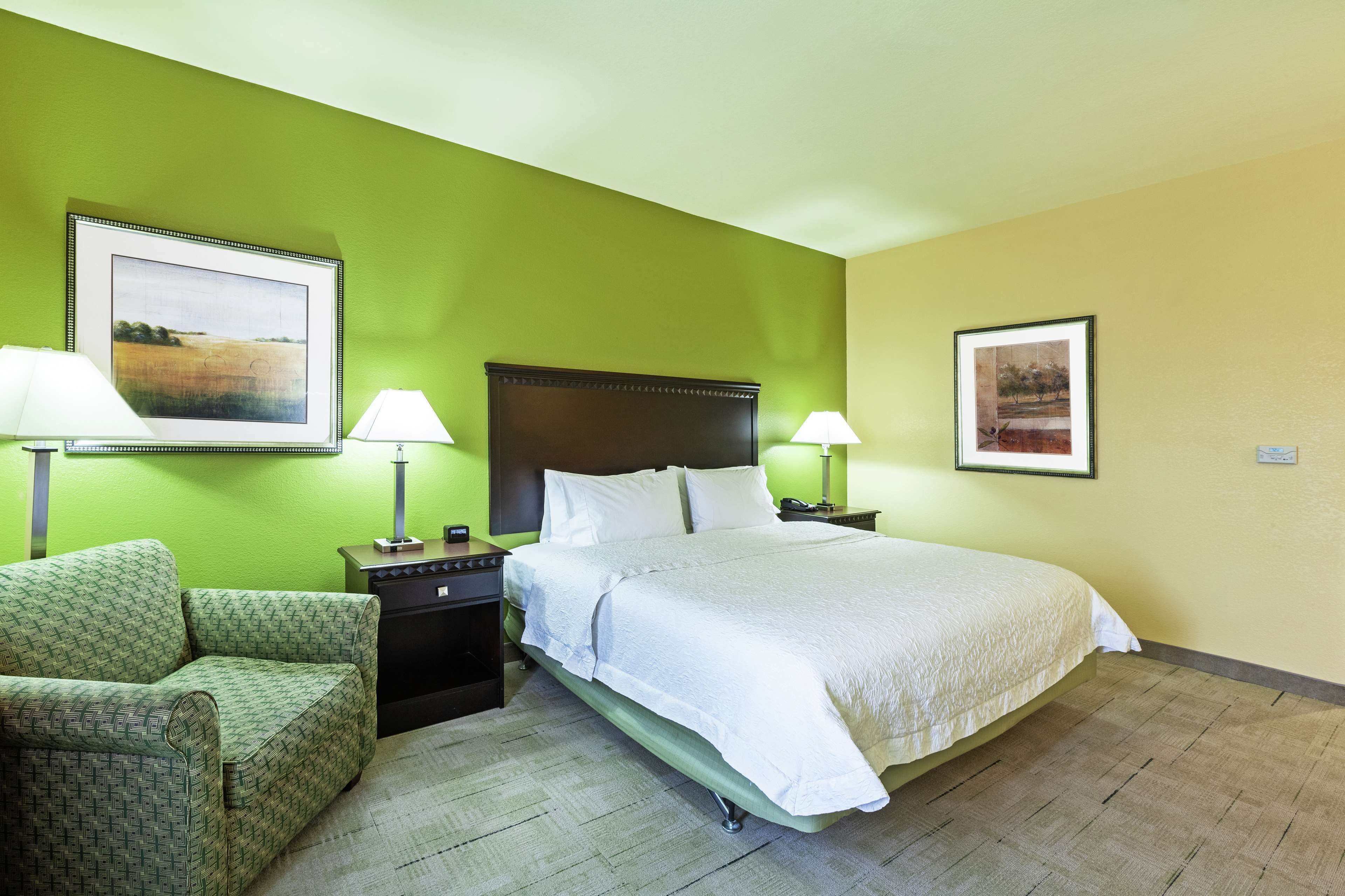 Una habitación de hotel con cama, dos mesitas de noche, una silla y dos lámparas de pared.