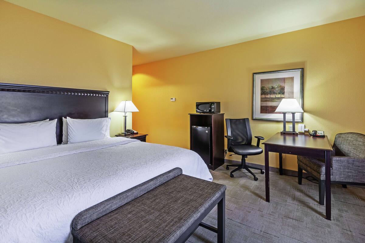 A hotel room with a large bed, bedside tables, a desk, a chair, a lamp, a microwave, and a framed picture on the wall.