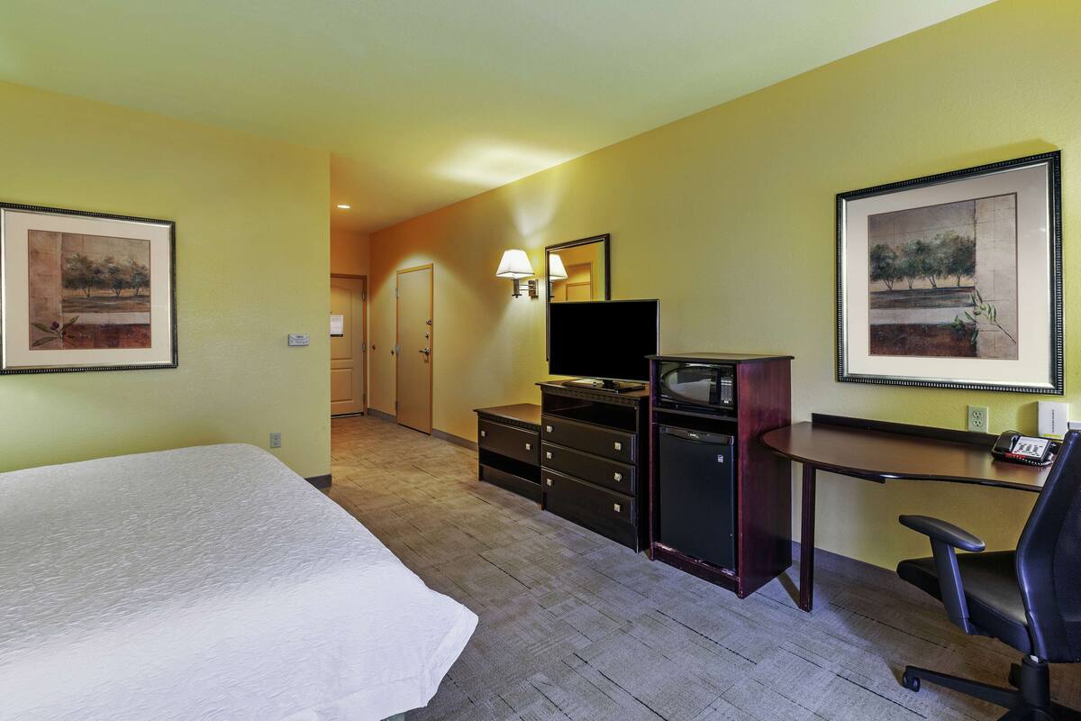 A hotel room with a bed, a desk with a chair, a television, and two framed pictures on the walls.