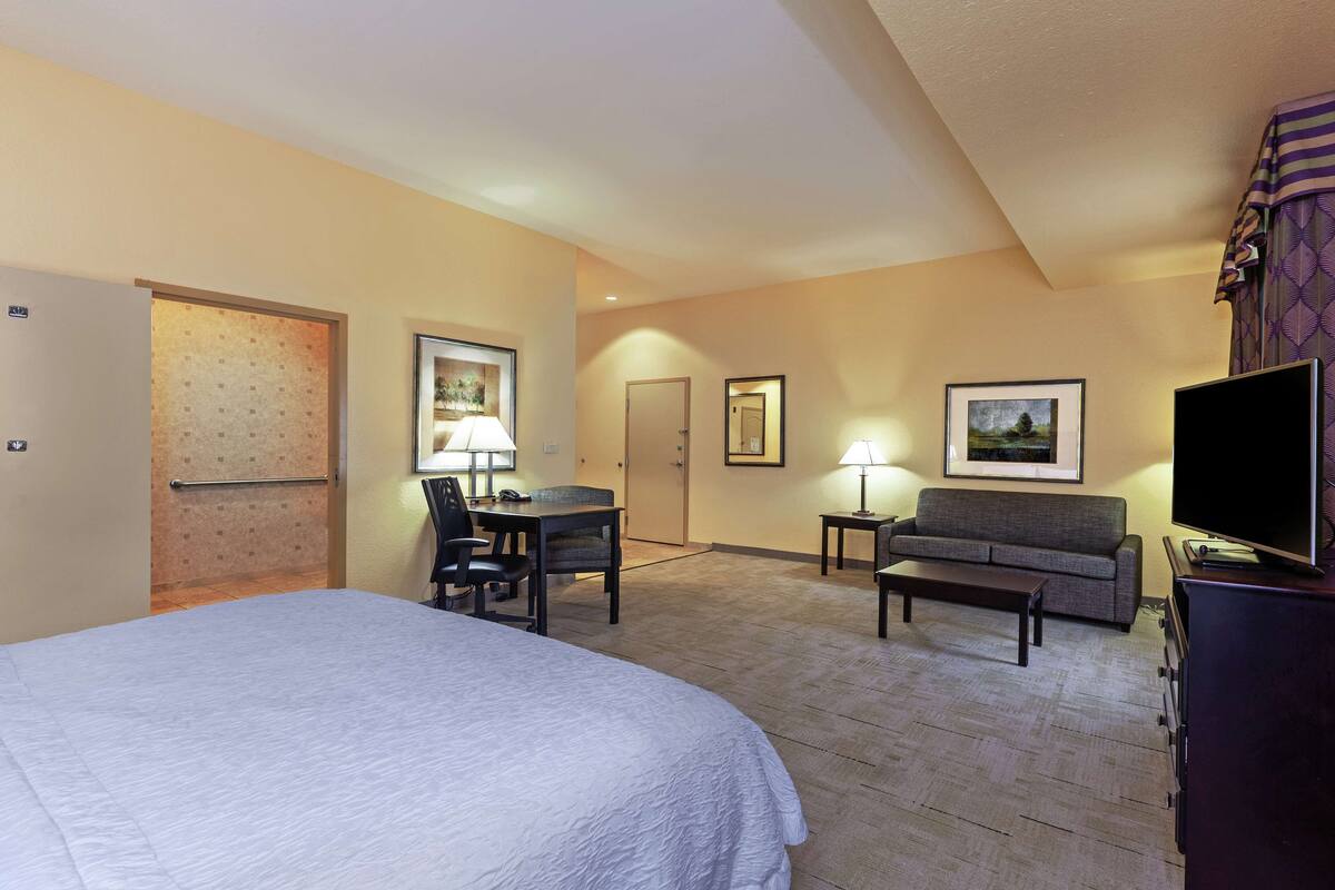 A hotel room with a large bed, a desk with a chair, a sofa, a TV, and framed pictures on the walls.