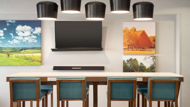 Un espace repas moderne avec une longue table et quatre chaises, un téléviseur à écran plat fixé au mur et trois lampes suspendues au-dessus.