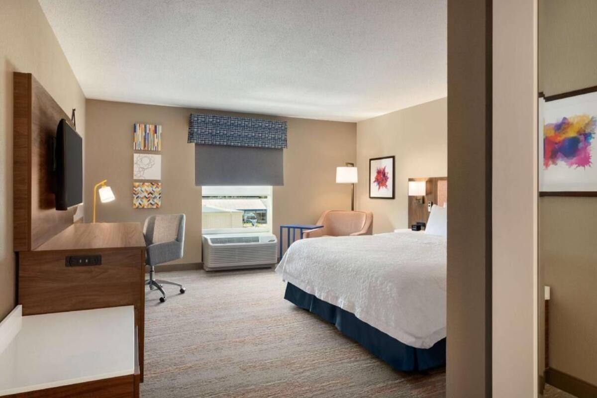 Une chambre d’hôtel équipée d’un lit, d’un bureau, d’une chaise, d’un climatiseur et décorée avec des œuvres d’art.