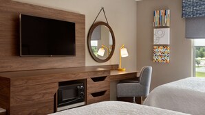 Une chambre d’hôtel équipée d’un téléviseur à écran plat, d’un miroir rond, d’un bureau avec une lampe, d’un four à micro-ondes et d’une chaise.