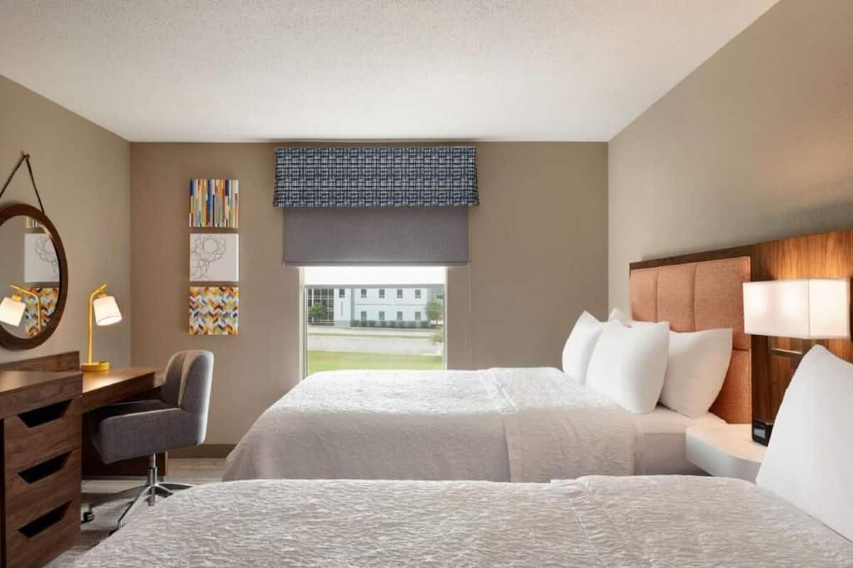 Une chambre d’hôtel avec deux lits, un bureau, une chaise, une grande fenêtre et des œuvres d’art accrochées aux murs.