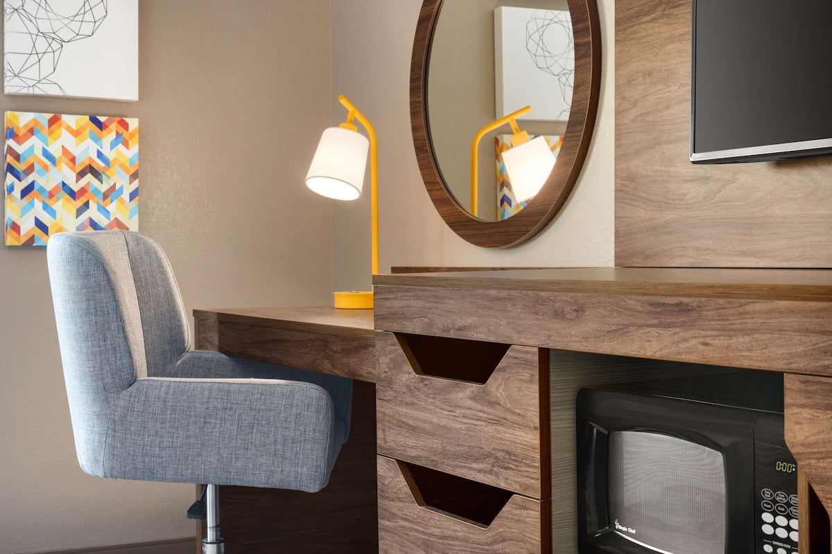 Une chambre d’hôtel moderne avec un bureau en bois, une chaise bleue, une lampe fixée au mur, un miroir et un téléviseur.