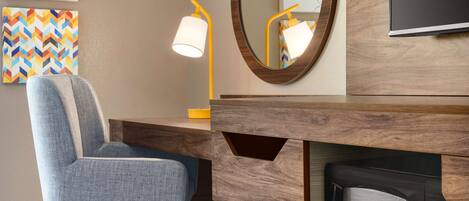A modern hotel room with a wooden desk, a blue chair, a wall-mounted lamp, a mirror, and a television.